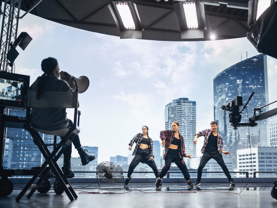 Three dancers perform in a studio with a cityscape backdrop. A person with a megaphone directs them.