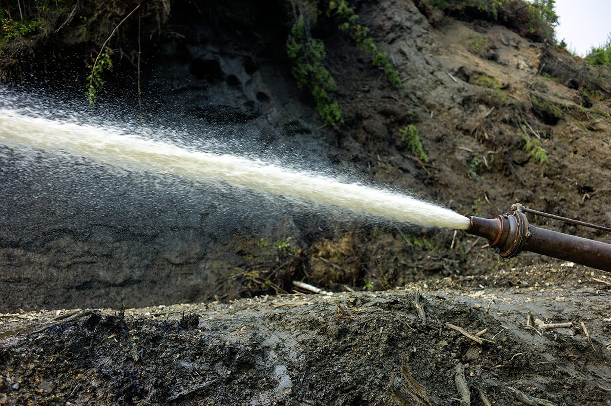 The giant monitor is spraying water onto the frozen muck and the permafrost is melting away at the Boneyard Alaska.. Placer mining for gold photo giant monitor water stream photo of the nozzle