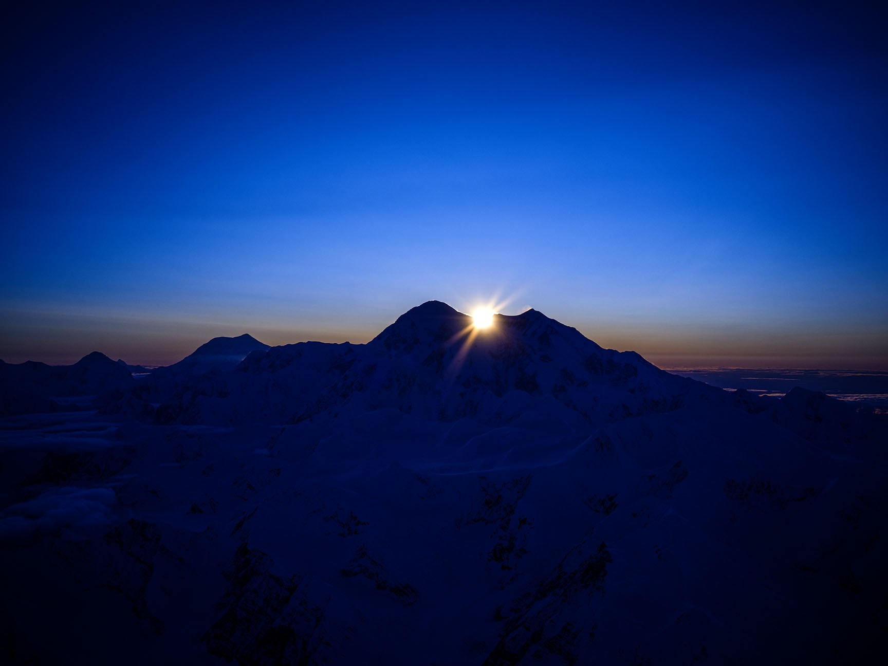 The sun is setting in the west, just above the very top of Mt Denali in Denali National Park Alaska. At 20,310 feet, Mount Denali is North Americas tallest mountain and quite the landmark. This aerial photo of the mountain show the South and the North peak and the elevation difference between the two as the wind is blowing snow off the north peak.