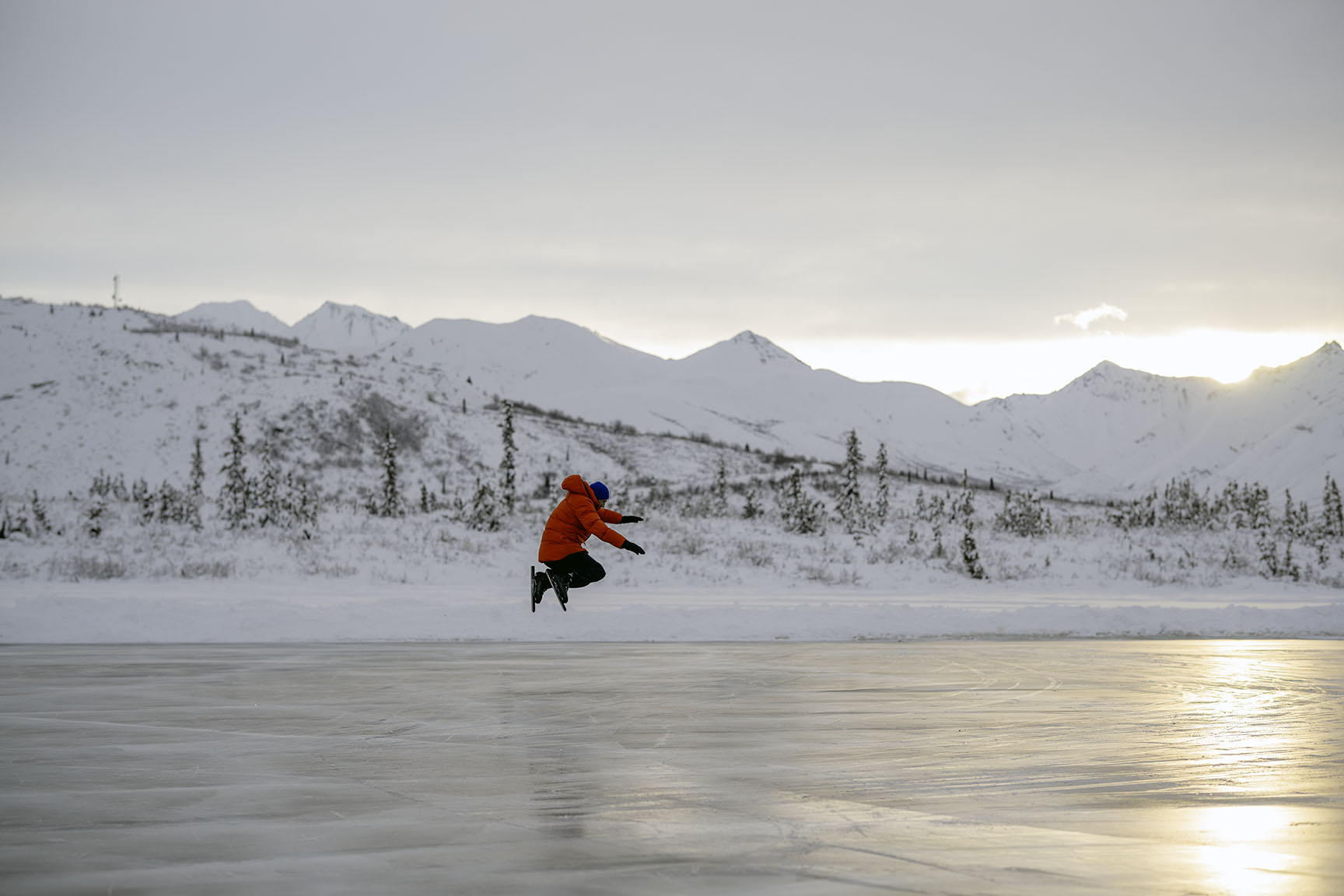 Video production shoot in Alaska, shooting a speed skater in an orange down jacket skating on the frozen lake, camera operator shooting from a low angle following the skater on the frozen lake, jumping for joy